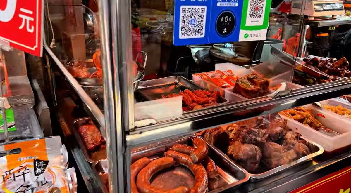 Trays of braised meats behind glass at a Xiamen street food stall, with QR payment signs above.